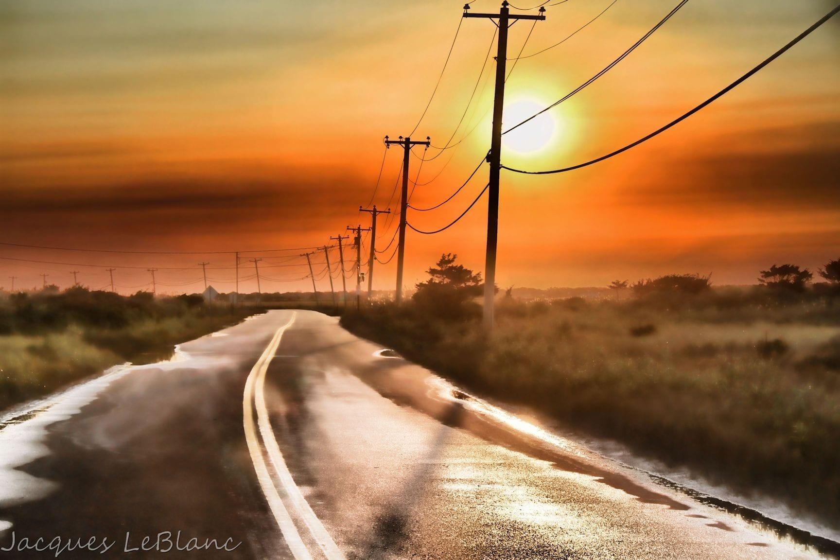A passing summer rainstorm has left Dune Road in Hampton Bays glistening and full of puddles and unveils a red sky sunset behind a line of telephone poles.